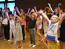 Eine große Gruppe von Kindern singt und tanzt mit erhobenen Händen in einem Probenraum.
