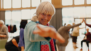 Eine Gruppe älterer Personen tanzt im Hintergrund, während eine Frau direkt auf die Kamera zutanzt.