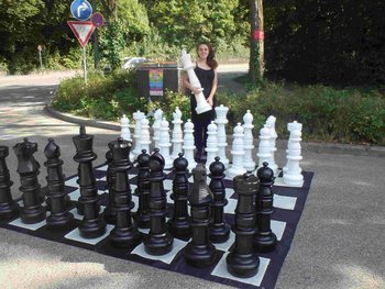 Eine junge Frau steht hinter einem großen Freiluftschachbrett und hält eine große Schachfigur in den Händen.
