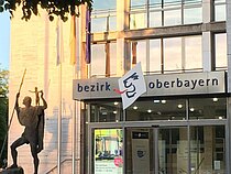 Der gläserne Haupteingang des Bezirks Oberbayern enthält links das große Ausstellungsplakat der Galerie und rechts einen Hebe-Lift. Neben dem Eingang befinden sich eine Christopherus Statue und drei Flaggen.