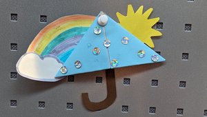 An einer Pinnwand wurde ein zum Regenschirm gestaltetes Papierlesezeichen befestigt. Der Schirm ist blau. An seiner linken Seite klebt eine weiße Wolke mit Regenbogen und an seiner rechten Seite eine gelbe Sonne.