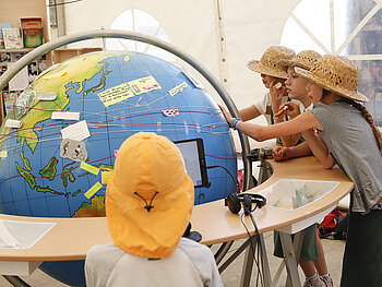 Mehrere Kinder stehen um einen Globus herum, der ungefähr so hoch ist wie sie. Auf dem Globus kleben handgeschriebene Zettel. 